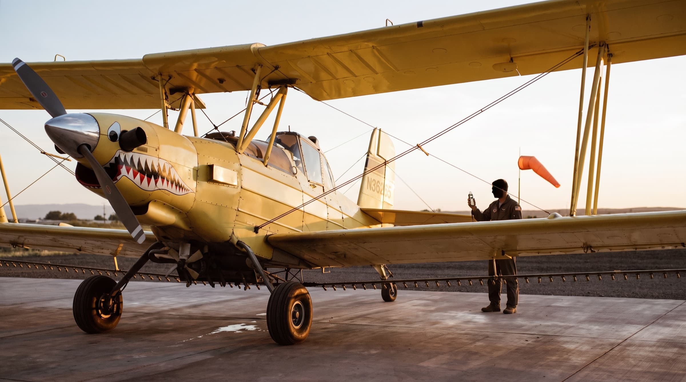 A yellow Ag Cat biplane parked by an orange windsock at the airstrip, with the sock half full in a light breeze while the pilot checks a handheld weather meter in calm morning light.
